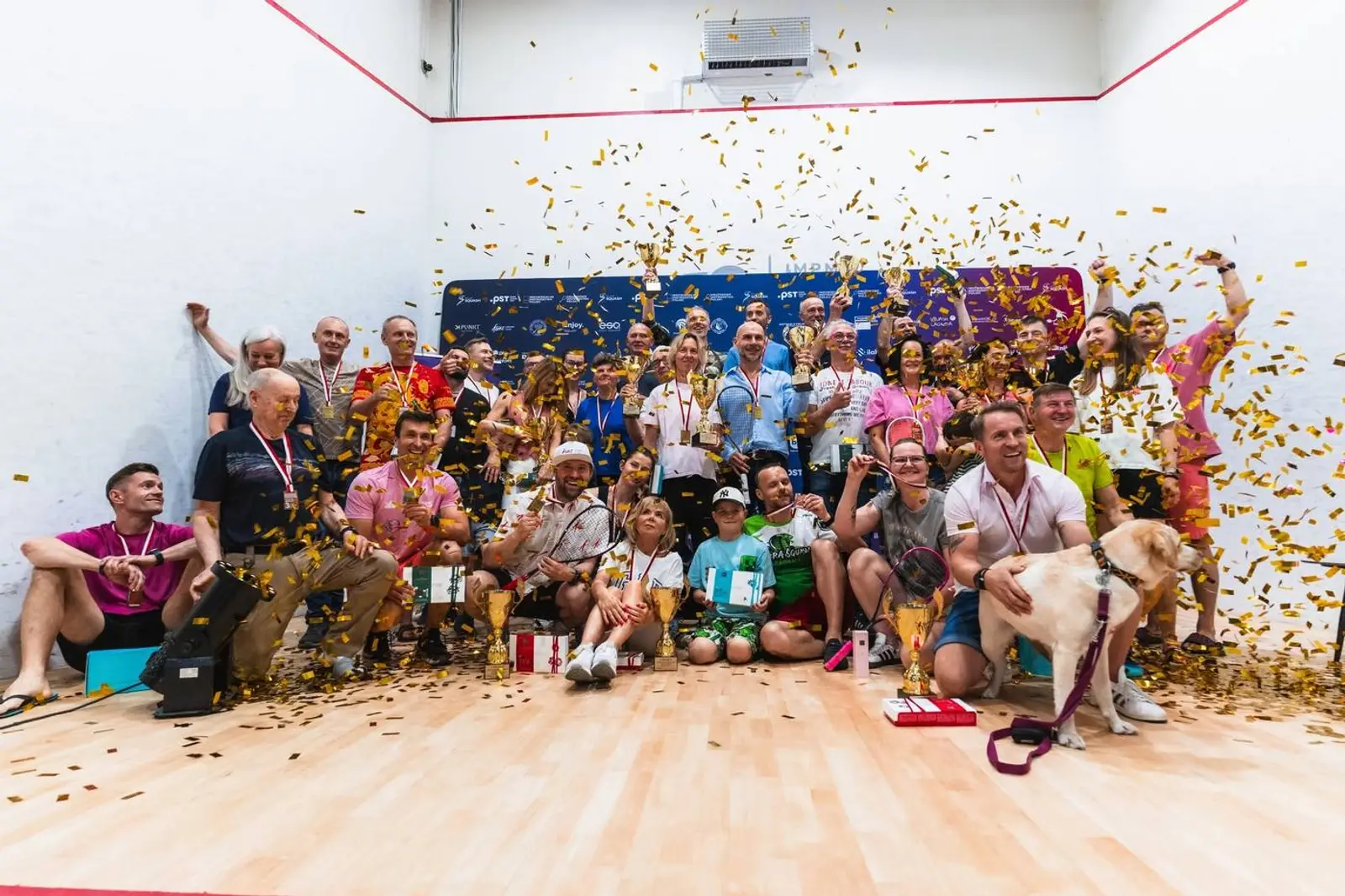 Roześmiana grupa uczestników 55+ z medalami na korcie squasha