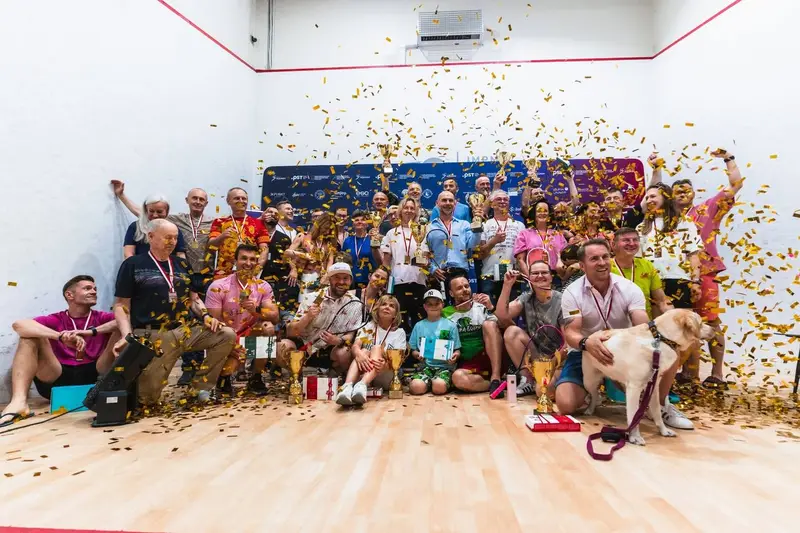 Roześmiana grupa uczestników 55+ z medalami na korcie squasha