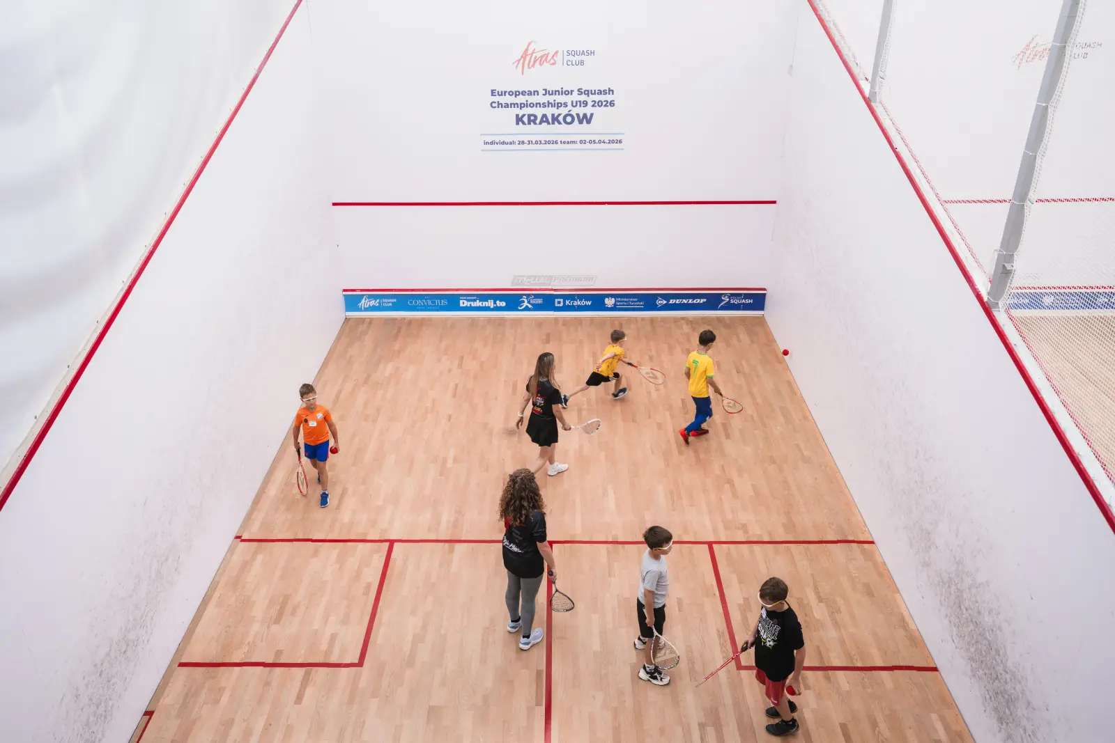 Dzieci z rakietami ćwiczą na szklanym korcie turniejowym Atras Squash Club