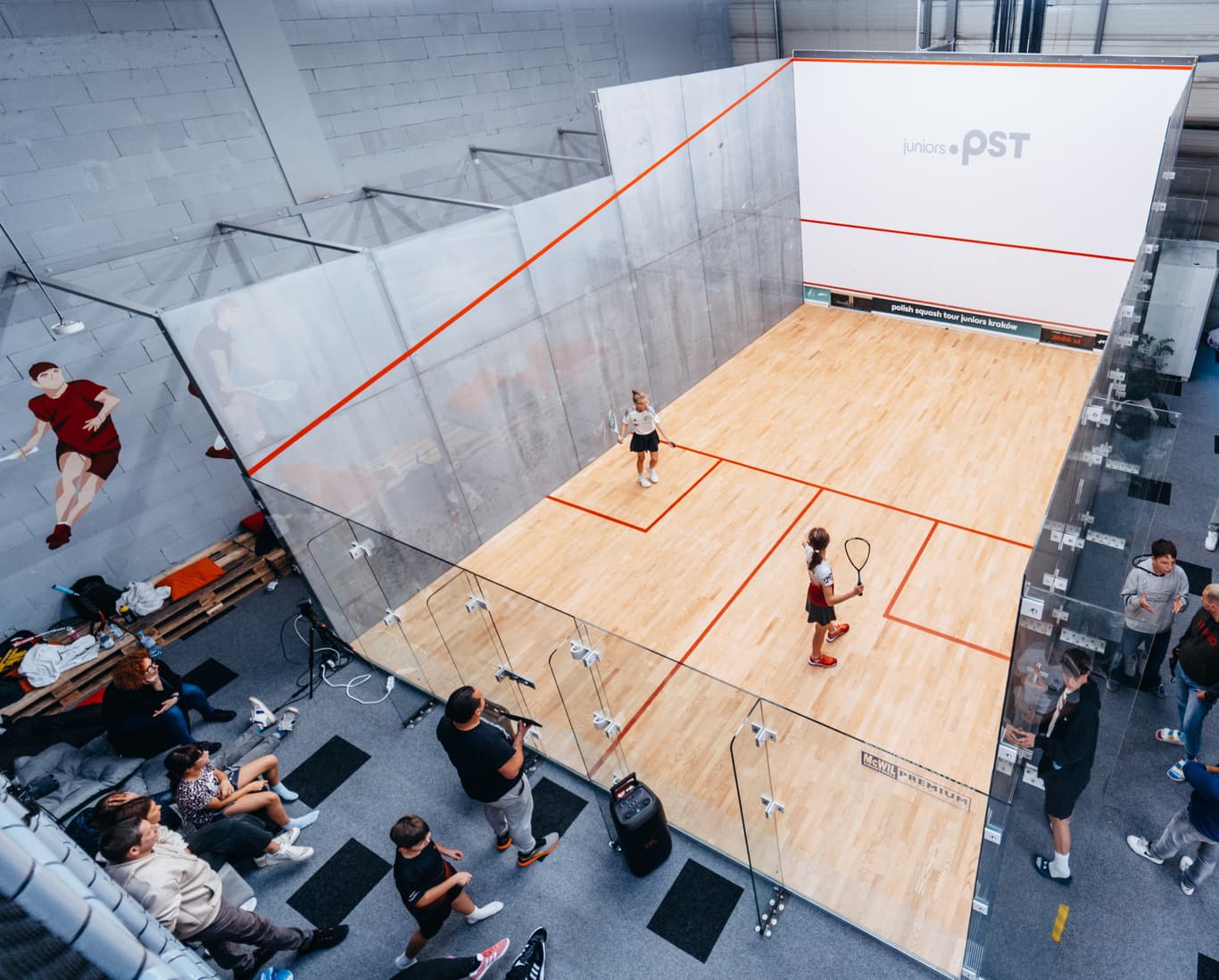 Aerial view of a glass-walled squash court with two players competing, while a small audience watches from outside the court.